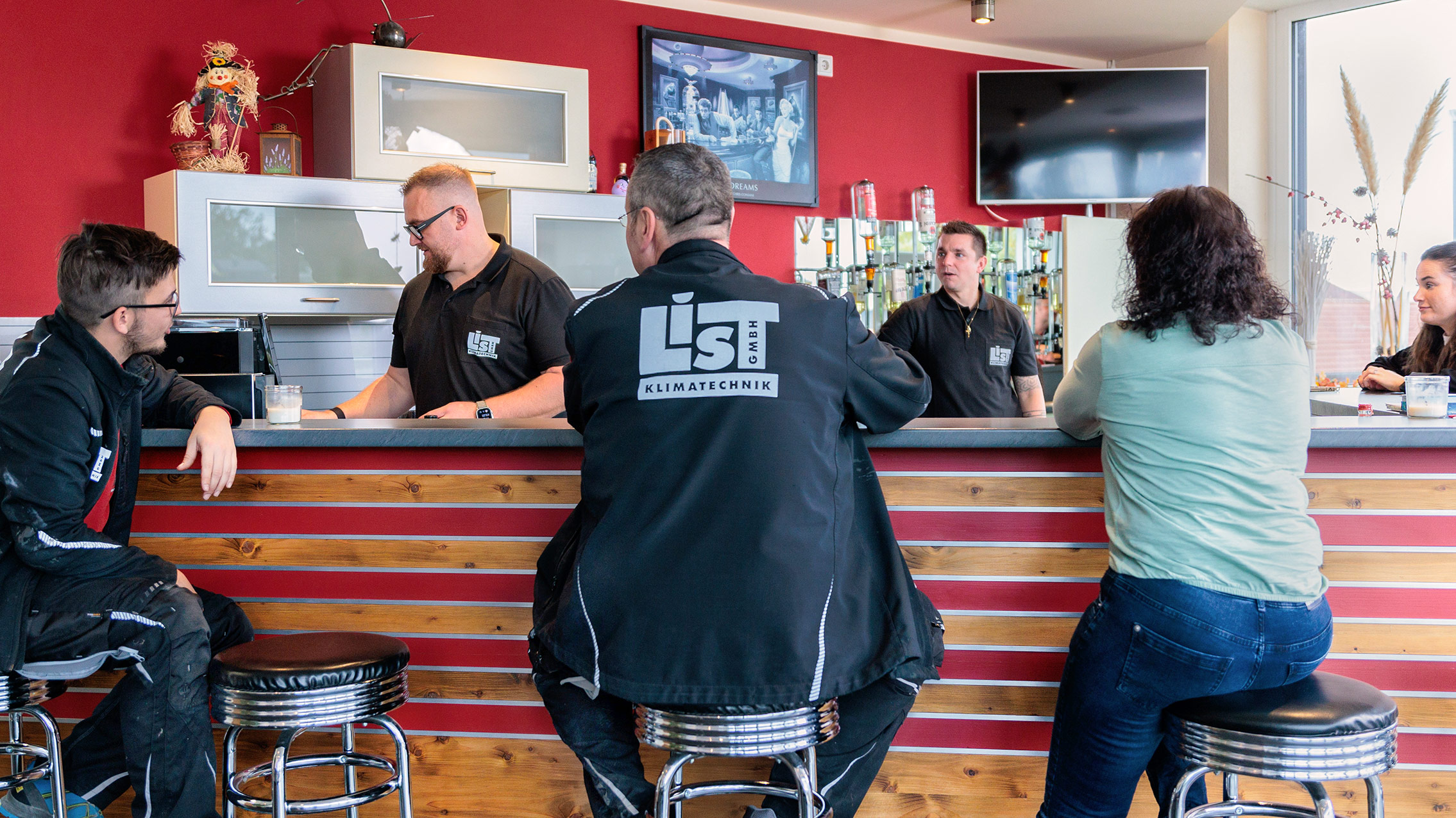 Das Team der Firma List trifft sich in der Küche zu einer entspannten Kaffeepause. Ein Mitarbeiter und eine Mitarbeiterin sitzen auf den Barstühlen vor der Theke. Florian List und ein weiterer Mitarbeiter stehen hinter der Bar an der Küchenzeile. Vivian List lehnt an der rechten Seite der Bar. Teamzusammenhalt wird groß geschrieben.