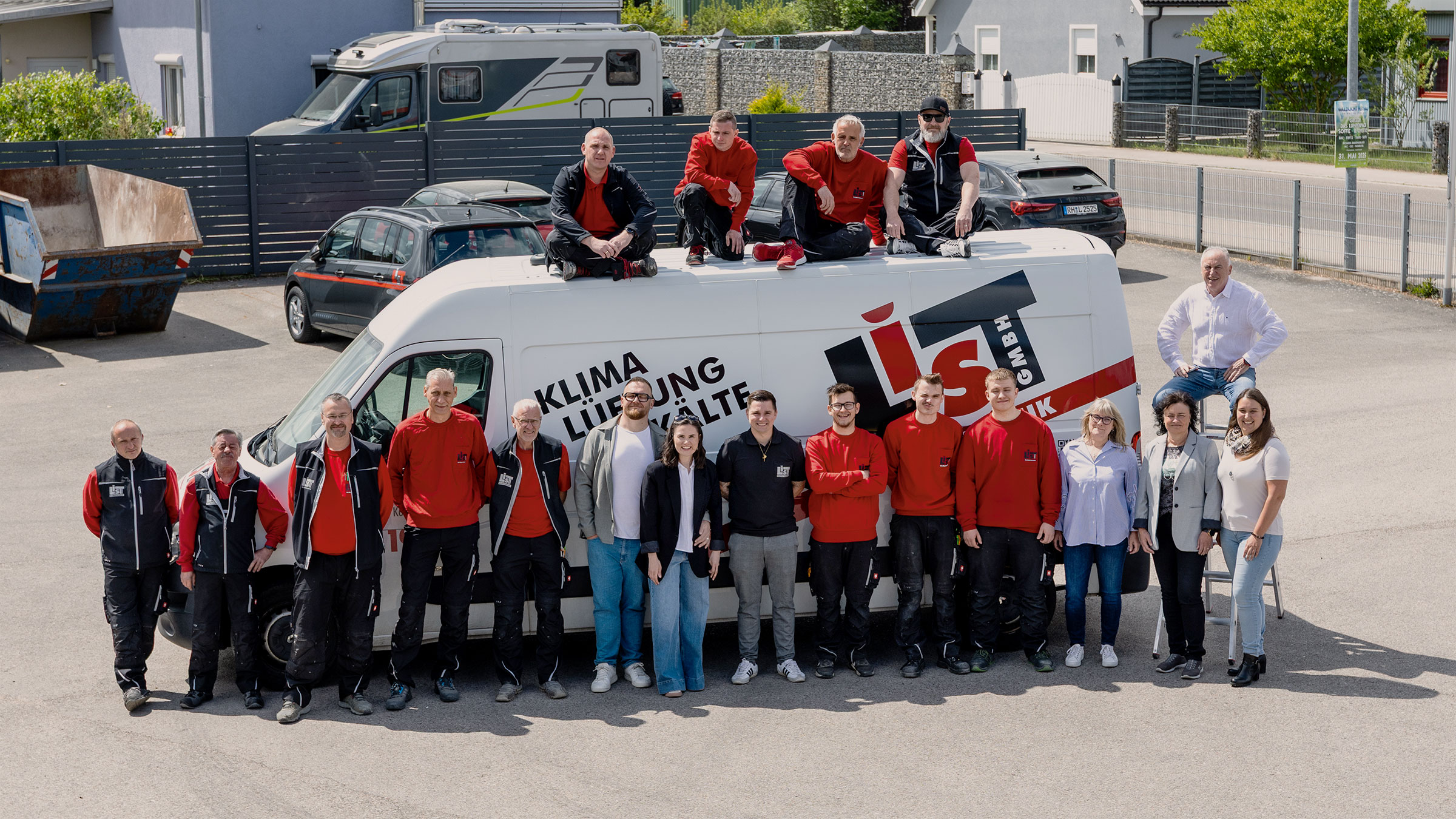Außenaufnahme im Hof des Unternehmens. Die Familie und die Mitarbeiter stehen vor einem Transporter. Ein Teil der Monteure ist auf dem Dach. Alle schauen in Richtung des Betrachters. Der Zusammenhalt des Familienunternehmens wird deutlich spürbar.
